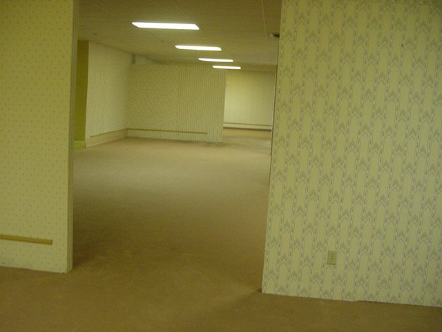 A large space with patterned wallpaer and beige floors and ceiling. Fluorescent panels provide muted lighting
